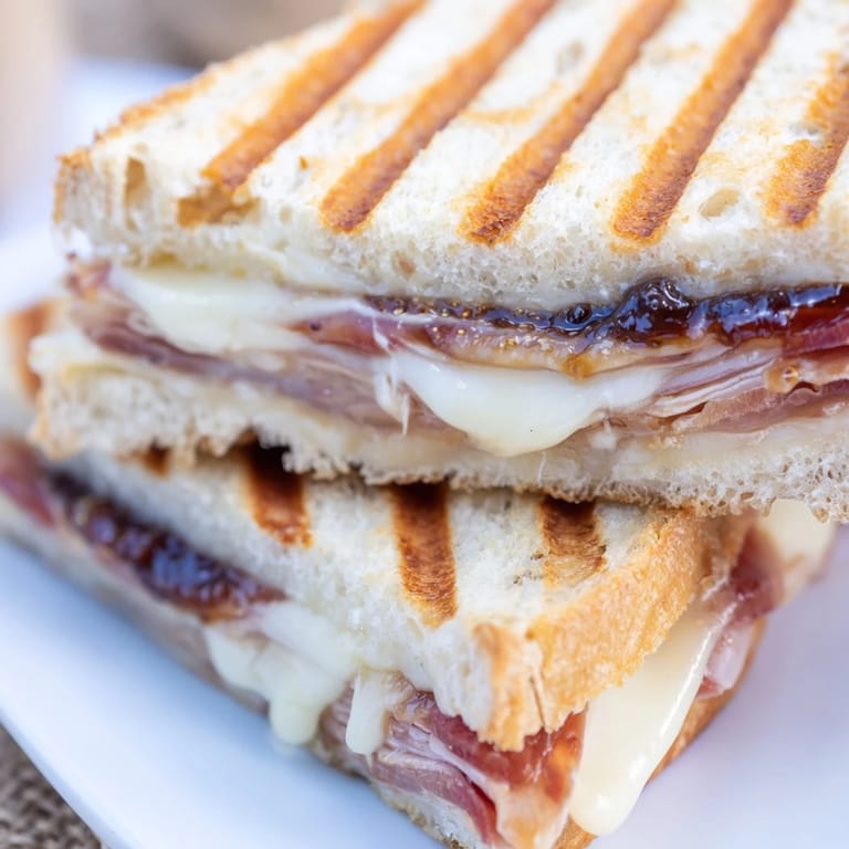 A toasted Crispy Prosciutto & Fig Panini rests on a wooden board, its crispy bread golden-brown and buttery, served with a side salad.